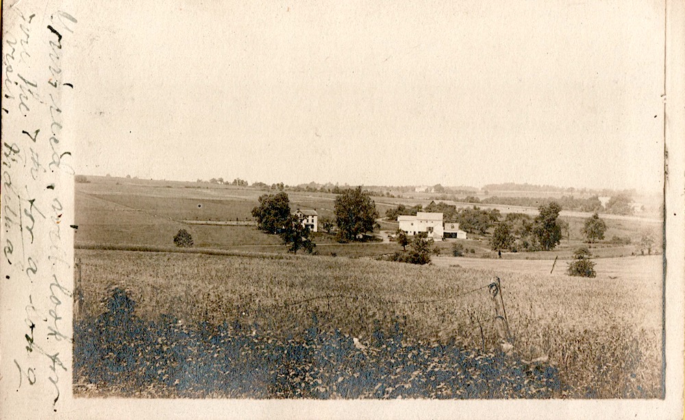 old black and white postcard has a handwritten note on the left-hand edge. The postcard image is a long view across a field of a farm in the distance.