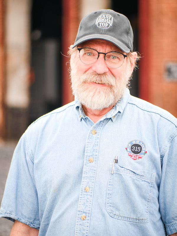 a man looks at the camera, wearing a hat that says East Broad top and a blue shirt with a logo for the Rio Grande line