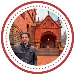 A man in a leather jacket stands in front of an ornate Victorian building
