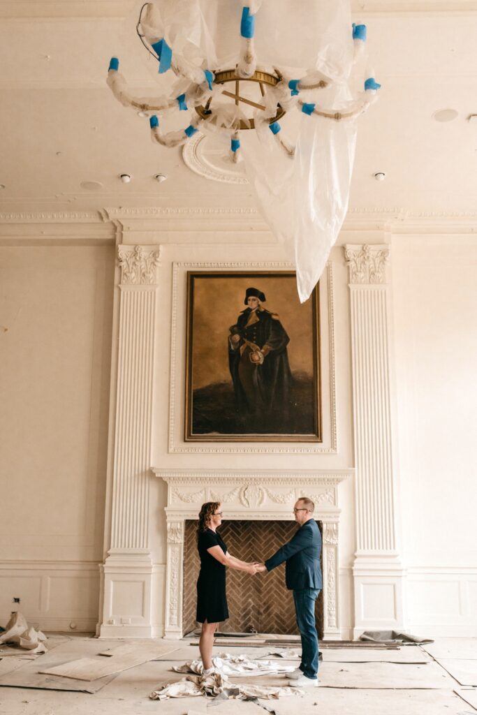 A couple hold hands in front of a fireplace with a monumental portrait hanging over it. They are in a room under construction with a large chandelier wrapped in protective covering. Soft daylight filters in from the right.