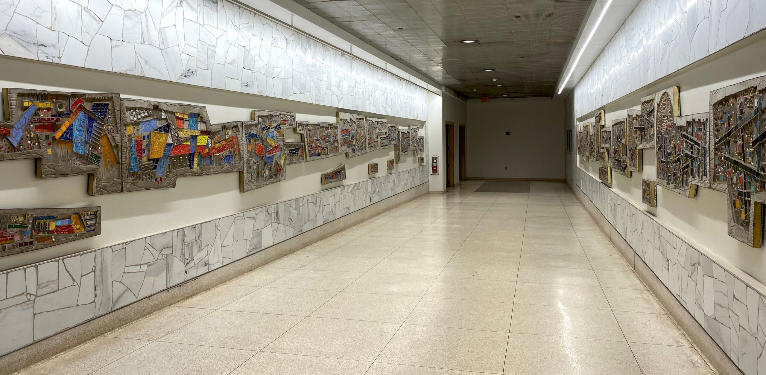 Long corridor displaying Virgil Cantini's colorful abstract mosaic panels mounted on both walls, with marble tile wainscoting and terrazzo floors at the Steel Plaza T Station, Pittsburgh